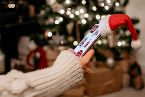 An arm in a sweater reaches out to scroll on a phone that has a tiny Santa Claus hat on it